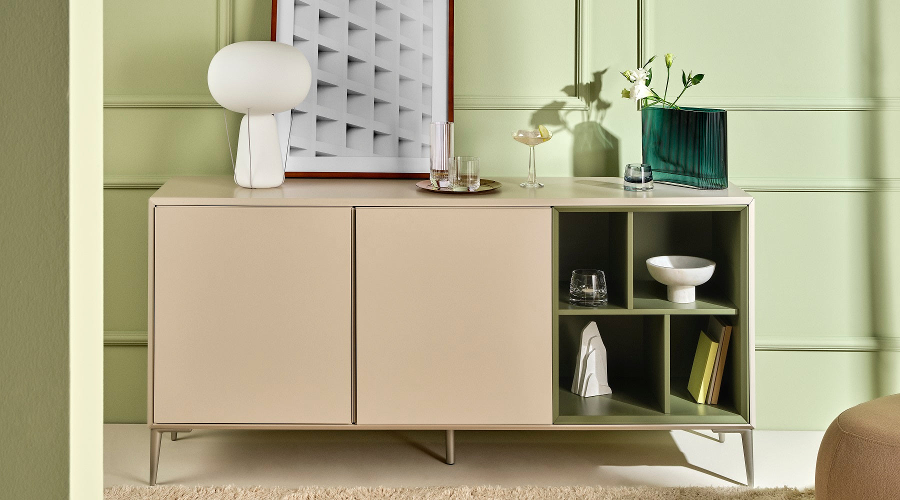 A home setting of a side supboard against a sage coloured wall, with various NUDE products; Mist vase wide in petroleum, the Big Top cocktail collection and the Joy votives in clear and steel blue