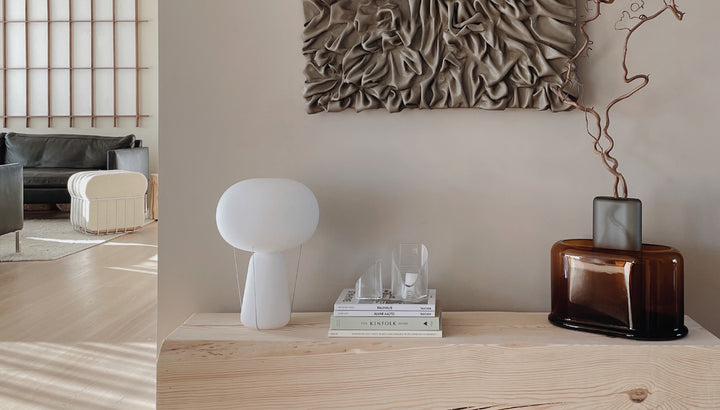 Japanda interior, wit a side table on which are presented the NUDE Blow lamp in opal white, a set of books on top of which are the Mist Lights votives in clear as well as the Layers vase wide in amber and Layers vase tall in grey with some brenches