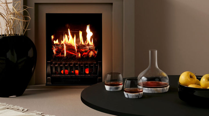 NUDE Chill carafe with marble base presented with 2 NUDE Chill whiskey glasses, in a wintery environment with a fireplace in the background