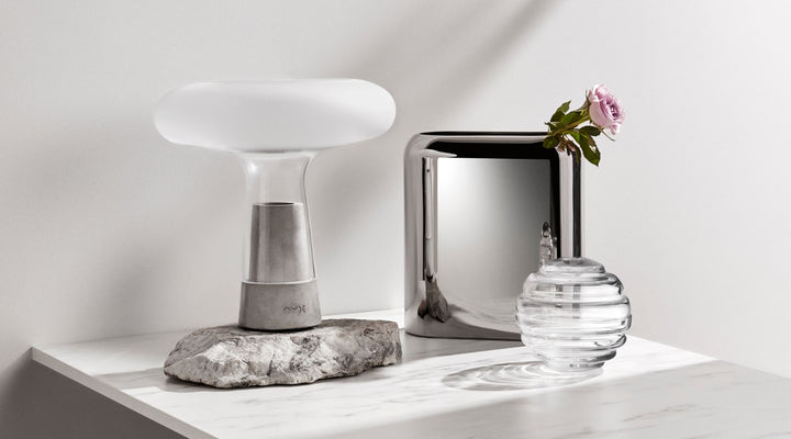 Corner of NUDE products on a marble table, the NUDE Orion tablelamp with frosted glass and cement foot, the Layers vase, a tall squareshaped vase in platinum coating and Nest, a candy box of round leadfree crystal with a relief of ribbles